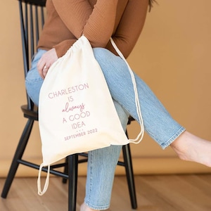 May include: A white drawstring bag with the text "Charleston is always a good idea... September 2022" printed on it. The bag is being held by a person wearing a brown sweater and blue jeans.