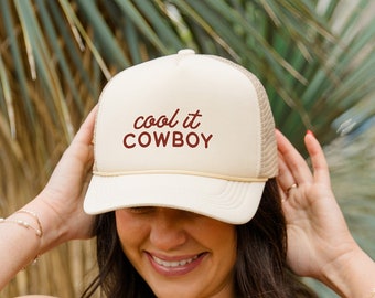 Könnte beinhalten: Eine Frau trägt eine beige Netz-Trucker-Kappe mit dem Text "cool it COWBOY" in Rot. Sie trägt ein schwarzes Bikini-Oberteil und eine blaue Jeans.