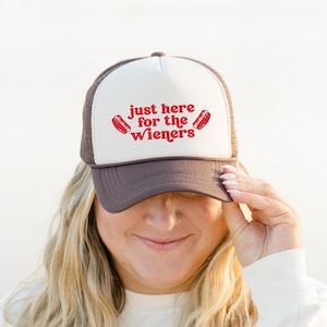 Op de afbeelding: Een bruine en witte truckerpet met de rode tekst "just here for the Wieners". De pet heeft een mesh achterkant en een effen voorpaneel. De tekst gaat vergezeld van twee rode hotdog illustraties.
