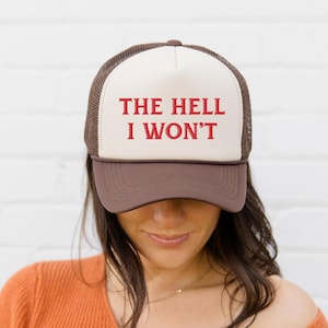 Puede incluir: Gorra de camionero con visera y parte trasera de malla marrón, panel frontal color crema y la frase "THE HELL I WON'T" en letras rojas. La gorra la lleva una persona con un jersey naranja.