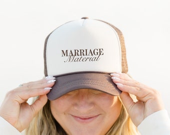 Puede incluir: Una mujer con una sudadera blanca sostiene una gorra de camionero marrón y blanca con el texto "Marriage Material" en letra marrón.