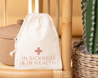 May include: A white cotton drawstring bag with a red cross and the text "IN SICKNESS & IN HEALTH" printed on it. The bag is sitting on a wooden shelf with books and a cactus plant.