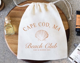 May include: A white drawstring bag with the text "CAPE COD, MA Beach Club Jake & Maddie 2024" printed on it. The bag is sitting on a wooden round board with a candle, matches, a small bottle of oil, a tube of hand lotion, and a tin of coffee.