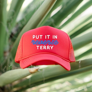 May include: Red trucker hat with a white mesh back. The hat has blue text that reads "PUT IT IN REVERSE TERRY".