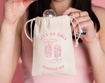 May include: A white drawstring bag with the text "Let's Go Girls Nashville 2023" and a pink outline of two cowboy boots. The bag is filled with silver disco balls.