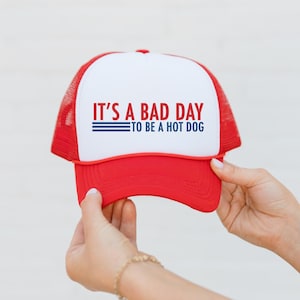 May include: A red and white trucker hat featuring the text "IT'S A BAD DAY TO BE A HOT DOG". The hat has a red peak and mesh back. A casual accessory for everyday wear.