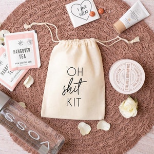 Può includere: Una borsa di tela bianca con il testo "OH SHIT KIT" su di essa. La borsa è piena di vari articoli, tra cui una bottiglia d'acqua, due bustine di tè, un piccolo barattolo di tartufi al champagne, un tubo di crema per le mani e un piccolo biglietto che dice "Mi sento bene". Gli articoli sono disposti su un tappetino intrecciato marrone.