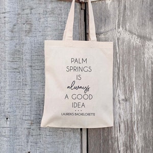 May include: A beige tote bag with black text that reads "Palm Springs is always a good idea... Lauren's Bachelorette". The bag is hanging on a wooden door.