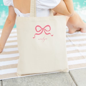 Puede incluir: Una bolsa de tela blanca con un lazo rosa y el nombre "evelyn" escrito debajo. La bolsa la lleva una persona que lleva una camisa blanca y un sombrero de paja que dice "bride".