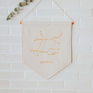 Könnte beinhalten: Ein beigefarbener Canvas-Banner mit einer goldenen Umrandung des Sternbilds Zwillinge und dem darunter gedruckten Wort "gemini". Der Banner hängt an einem dekorativen Haken an einer weißen Backsteinwand.