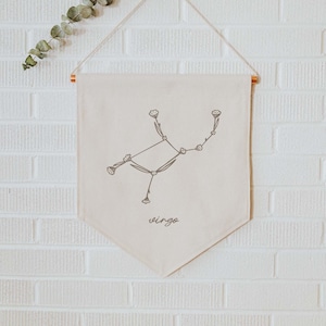 Könnte beinhalten: Ein beigefarbener Canvas-Banner mit einem floralen Sternbild-Design, das das Sternzeichen Jungfrau darstellt. Der Banner hängt an einem dekorativen Haken an einer weißen Backsteinwand. Der Banner trägt den Schriftzug "Virgo" in Kursivschrift am unteren Rand.