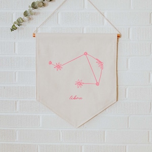 Könnte beinhalten: Eine weiße Leinwand-Tapete mit einer rosa Umrandung der Sternbilder Waage und dem Wort "libra" darunter. Die Tapete hängt an einem Haken an einer weißen Backsteinwand.
