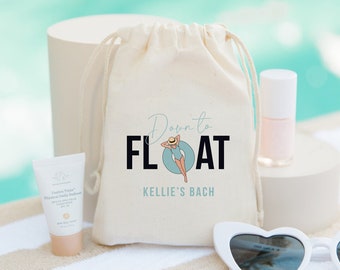 May include: A white canvas bag with a drawstring closure. The bag features a graphic of a woman in a swimsuit floating on a pool float with the text "Down to FLOAT" and "Kellie's Bach" printed on it. The bag is placed on a white and brown striped towel alongside a small bottle of nail polish, a tube of sunscreen, and a pair of white heart-shaped sunglasses.