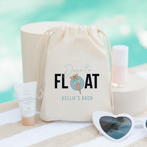 Puede incluir: Una bolsa de lona blanca con cierre de cordón. La bolsa tiene un gráfico de una mujer en traje de baño flotando en un flotador de piscina con el texto "Down to FLOAT" y "Kellie's Bach" impreso en ella. La bolsa está sentada sobre una toalla de rayas blancas y marrones con una pequeña botella de esmalte de uñas, un tubo de protector solar y un par de gafas de sol blancas en forma de corazón.