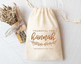 May include: A small white fabric bag with a drawstring closure. The bag has a brown print that says "Thankful for Hannah Friendsgiving 2022".