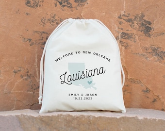 May include: White drawstring bag with the text "Welcome to New Orleans, Louisiana" and a blue outline of the state of Louisiana with a small heart in the center. The text "Emily & Jason 10.22.2022" is printed below the state outline.