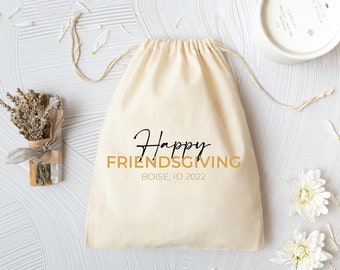 May include: A beige drawstring fabric pouch featuring the black and gold text Happy FRIENDSGIVING BOISE, ID 2022. Beside the pouch are a small bundle of dried lavender and herbs tied with twine, a white candle in a ceramic jar, and white flower petals.