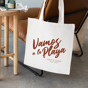 May include: A white canvas tote bag with brown handles. The bag has the text "Vamos a la Playa" in brown script lettering. Below the text is "Maddy & Conner 8.27.2023" in smaller brown lettering.