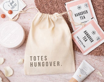 May include: A white canvas bag with the text "TOTES HUNGOVER." printed on it. The bag is filled with a variety of items, including a small box of chocolate truffles, two tea bags, and a tube of face cream. The tea bags are pink and white and have the text "HANGOVER TEA" printed on them. The face cream is in a white tube with a pink label. The items are arranged on a pink and white surface with white rose petals scattered around.