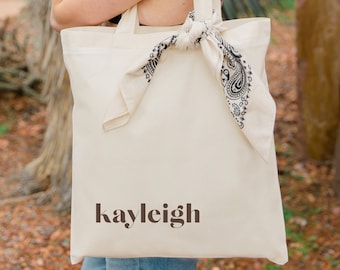 Puede incluir: Una bolsa de lona blanca con un pañuelo negro atado alrededor de las asas. La bolsa tiene el nombre "kayleigh" impreso en la parte delantera en color marrón.