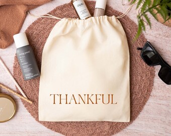 May include: A cream-colored drawstring pouch with the word THANKFUL printed in brown letters. The pouch is surrounded by a pair of brown socks, a Ouai hair oil bottle, a Living Proof dry shampoo bottle, a pink facial roller, and a candle.