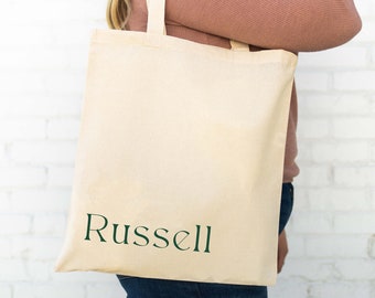Puede incluir: Una persona sostiene una bolsa de lona beige con la palabra "Russell" impresa en verde en la parte delantera.