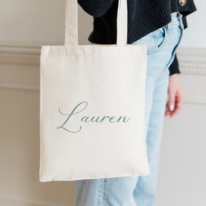 Puede incluir: Una bolsa de lona blanca con el nombre "Lauren" impreso en letra cursiva verde.