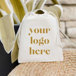 以下が含まれることがあります： 「your logo here」のゴールドテキストが入った、ナチュラルキャンバスの巾着袋。バッグは織り込まれた表面に置かれ、背景には緑と白の葉があります。ブランディングやプロモーションに最適です。