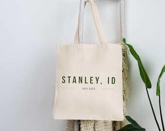 Puede incluir: Una bolsa de lona beige con el texto "STANLEY, ID" y "JULIO 2023" impreso en verde. La bolsa está colgada de una escalera de madera blanca.