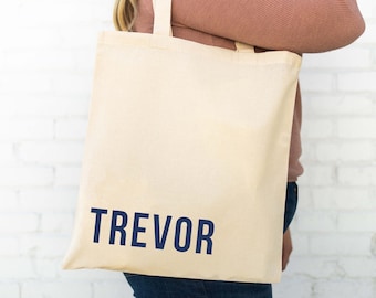 Puede incluir: Una bolsa de lona beige con asas azul marino y el nombre "TREVOR" impreso en azul marino en la parte delantera.