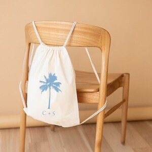 May include: A white drawstring bag with a blue palm tree design and the initials "C+S" printed on the front. The bag is hanging on the back of a wooden chair.