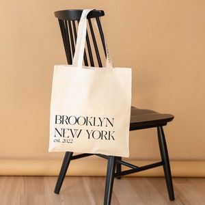 Op de afbeelding: Een witte canvas draagtas met zwarte handvatten en de tekst "BROOKLYN NEW YORK est. 2022" gedrukt op de voorkant. De tas hangt aan de rugleuning van een zwarte houten stoel.