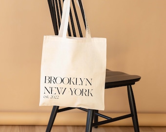 May include: A white canvas tote bag with black handles and the text "BROOKLYN NEW YORK est. 2022" printed on the front. The bag is hanging on the back of a black wooden chair.