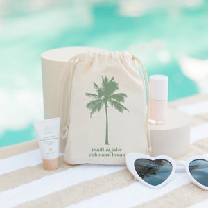May include: A white canvas bag with a green palm tree design and the text "madi & jake cabo san lucas". The bag is next to a white heart-shaped sunglasses, a small bottle of pink nail polish, and a small tube of sunscreen.
