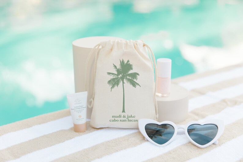 May include: A white canvas bag with a green palm tree design and the text "madi & jake cabo san lucas". The bag is next to a white heart-shaped sunglasses, a small bottle of pink nail polish, and a small tube of sunscreen.