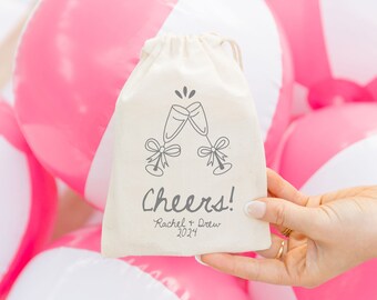 May include: A white cotton drawstring bag with a grey illustration of two champagne glasses clinking. The bag has the text "Cheers! Rachel & Drew 2024" printed on it.