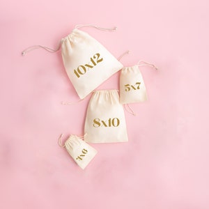 May include: Four white cotton drawstring bags with gold lettering on a pink background. The bags are labeled with different sizes: 10x12, 8x10, 5x7, and 6x8.