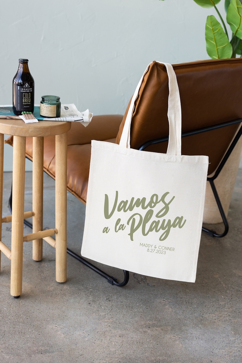 K&ouml;nnte beinhalten: Eine wei&szlig;e Canvas-Tragetasche mit dem Text "Vamos a la Playa" in gr&uuml;ner Schrift. Unter dem Text steht "Maddy & Conner 8.27.2023" in schwarzer Schrift.