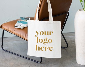 Puede incluir: Una bolsa de lona blanca con asas marrones colgando de una silla de cuero marrón. La bolsa tiene el texto "your logo here" impreso en dorado en la parte delantera.