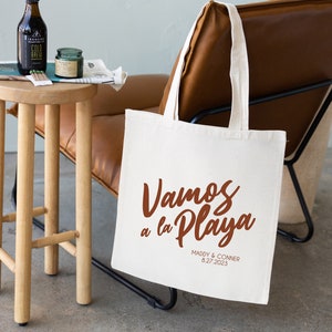 K&ouml;nnte beinhalten: Wei&szlig;e Canvas-Tragetasche mit braunen Henkeln. Die Tasche tr&auml;gt den Text "Vamos a la Playa" in brauner Schrift. Unter dem Text steht "Maddy & Conner 8.27.2023" in kleinerer brauner Schrift.