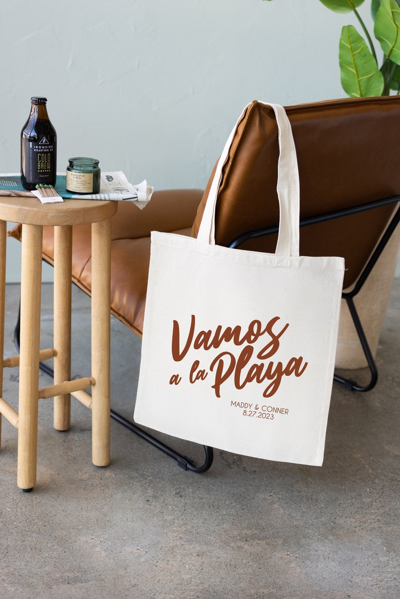 K&ouml;nnte beinhalten: Wei&szlig;e Canvas-Tragetasche mit braunen Henkeln. Die Tasche tr&auml;gt den Text "Vamos a la Playa" in brauner Schrift. Unter dem Text steht "Maddy & Conner 8.27.2023" in kleinerer brauner Schrift.