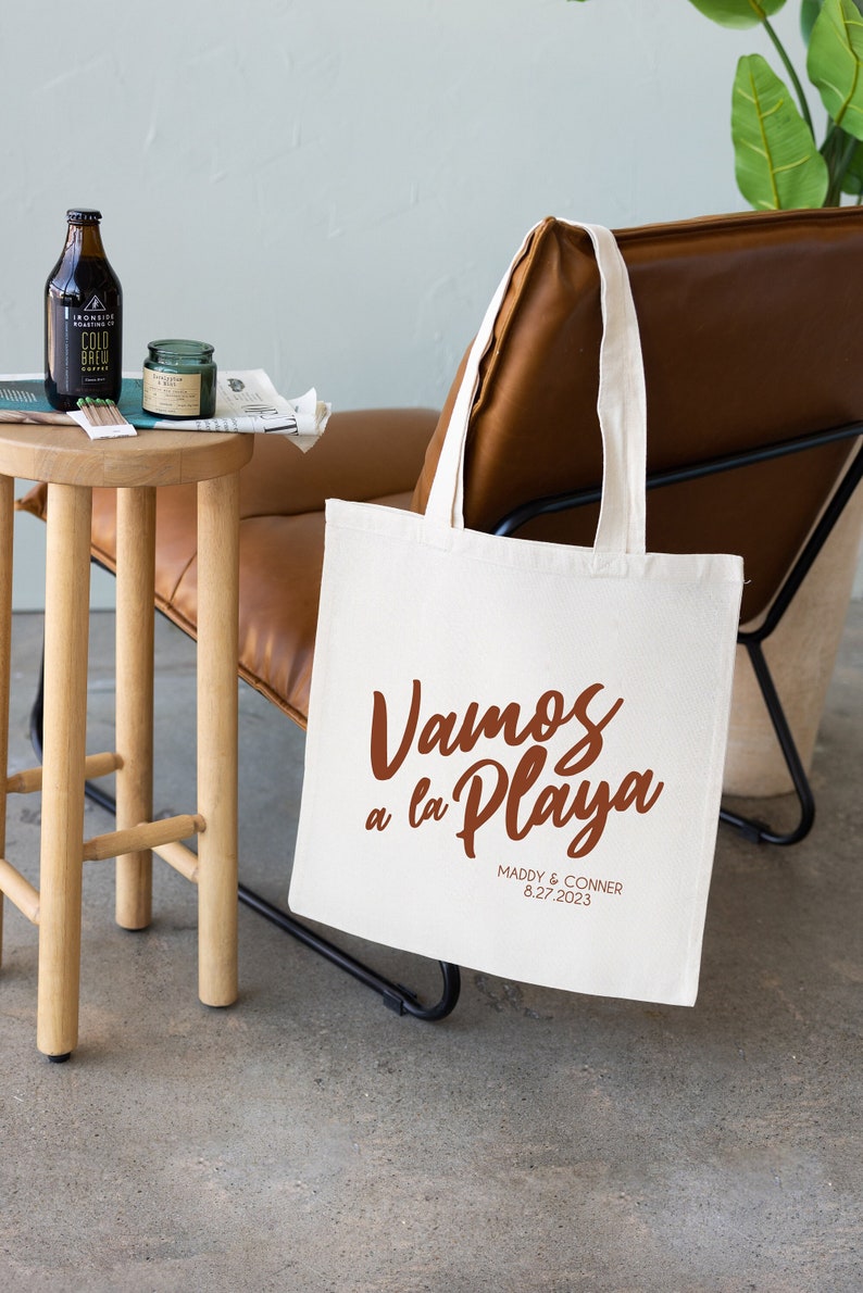 May include: A white canvas tote bag with brown handles. The bag has the text "Vamos a la Playa" in brown script lettering. Below the text is "Maddy & Conner 8.27.2023" in smaller brown lettering.