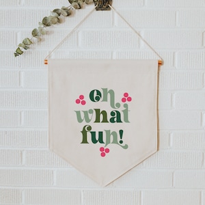 Op de afbeelding: Een witte canvasbanner met de tekst "oh what fun!" in groene en roze letters. De banner hangt aan een decoratieve haak aan een witte bakstenen muur.