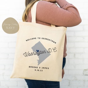 May include: Cream-colored tote bag with the text "WELCOME TO GEORGETOWN Washington D.C." in navy blue. A gray outline of the Georgetown area is in the center, with a navy blue heart. The names "BROOKE & JAMES" and the date "9.10.23" are printed below.