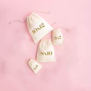 May include: Four white cotton drawstring bags with gold lettering on a pink background. The bags are labeled with different sizes: 10x12, 8x10, 5x7, and 6x8.