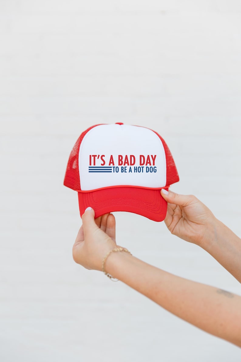 May include: A red and white trucker hat featuring the text "IT'S A BAD DAY TO BE A HOT DOG". The hat has a red peak and mesh back. A casual accessory for everyday wear.