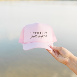 May include: Pink trucker hat with a brown embroidered design that says "LITERALLY just a girl".