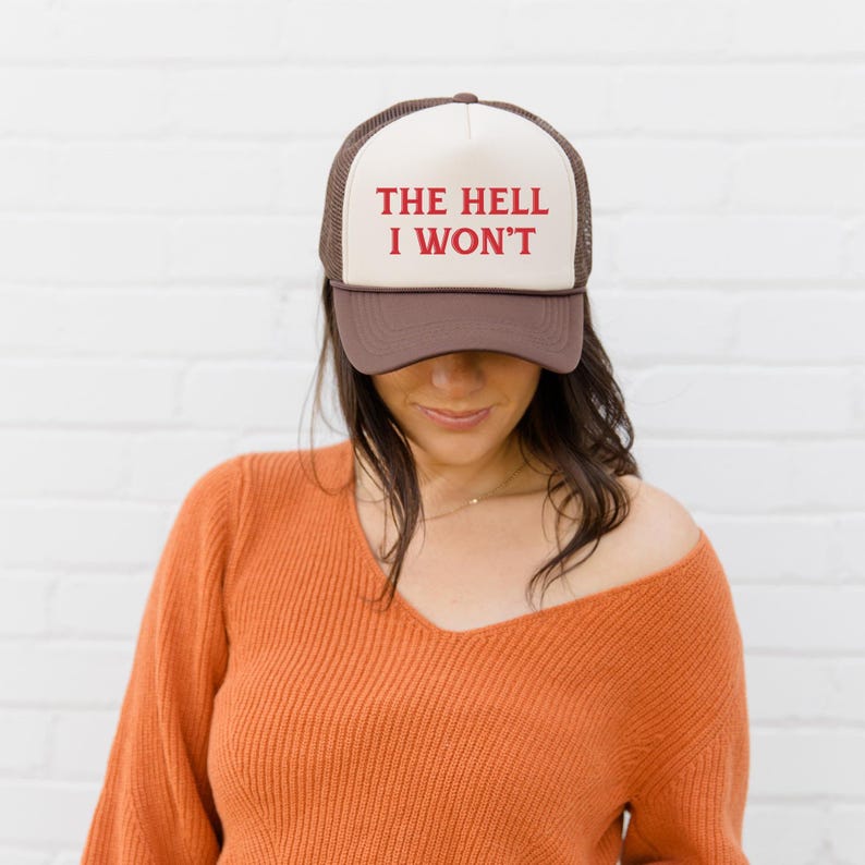 May include: A trucker hat with a brown brim and mesh back, a cream-colored front panel, and the phrase "THE HELL I WON'T" in red lettering. The hat is worn by a person wearing an orange sweater.