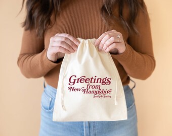 May include: A white drawstring bag with the text "Greetings from New Hampshire" in maroon lettering. The text "Smith & Jordan" is printed in smaller maroon lettering below the main text.