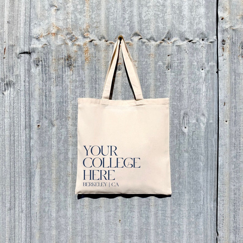 Puede incluir: Una bolsa de lona beige con el texto "YOUR COLLEGE HERE BERKELEY | CA" impreso en azul oscuro.